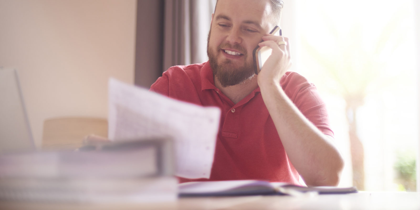 Bearded man in a red shirt sitting at a table, talking on a smartphone, and holding a piece of paper