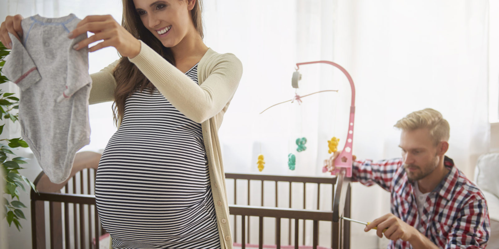 pregnant woman holds up a onesie while man in the background assembles wooden baby crib