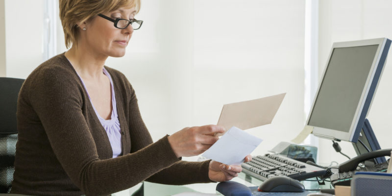 woman in brown sweater and black glasses sits at a computer and looks at papers in her hands