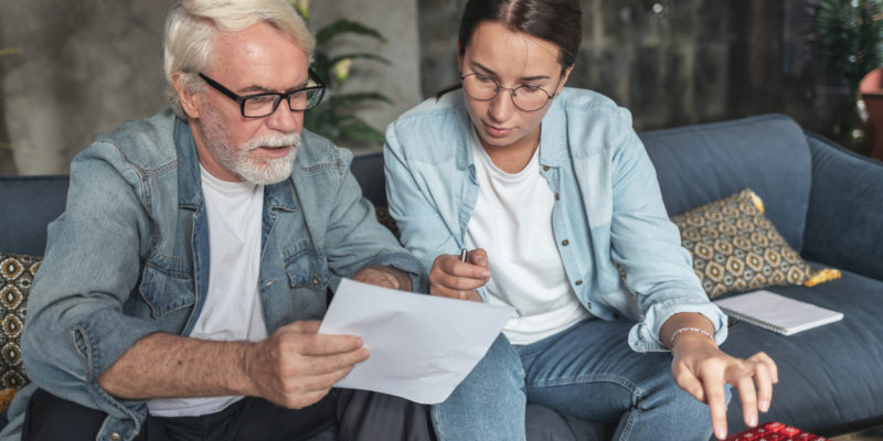 older man in glasses and jean shirt holds a piece of paper and talks while younger girl in glasses and jean shirt pushes buttons on calculator
