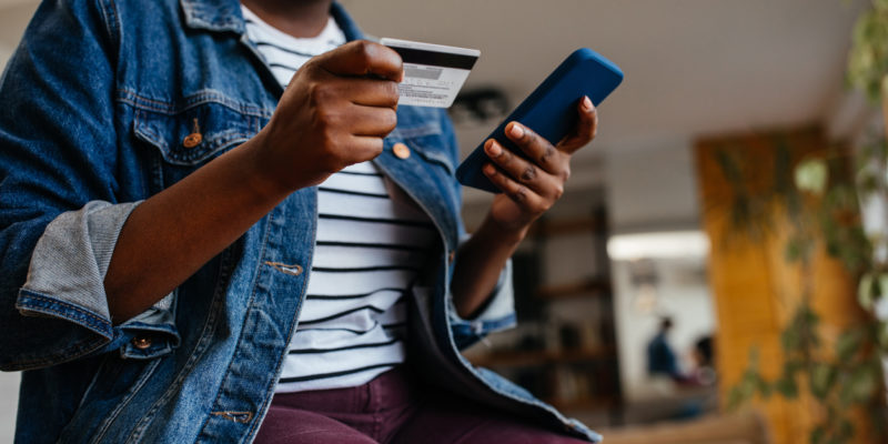 woman in a white striped shirt and jean jacket holds a credit card and uses a smart phone