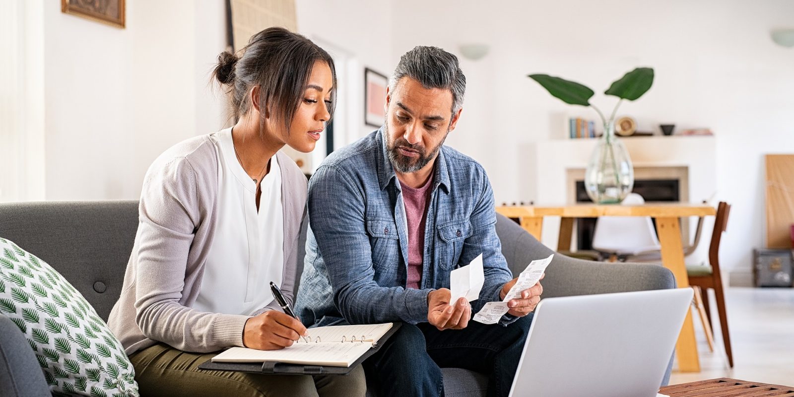 man holding receipts and woman writing in notebook sit on a couch in front of a laptop