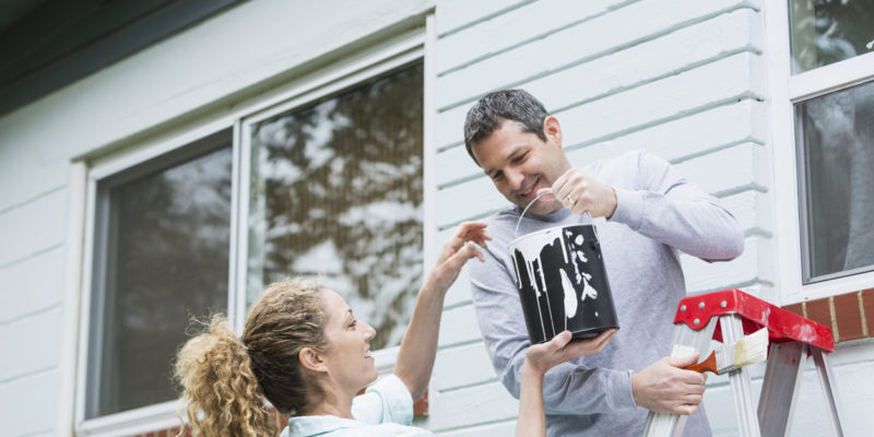 man on a ladder in front of a house accepts bucket of white paint from a curly-haired woman