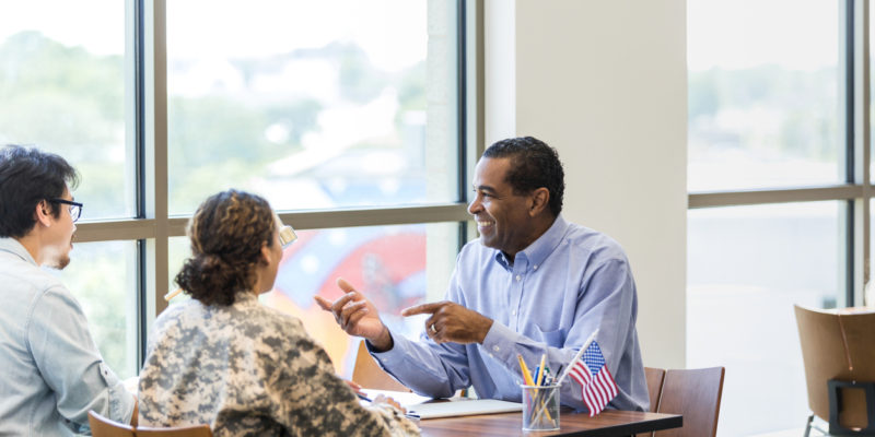 woman in camo and a man with glasses sitting at a table and talking with a man in a blue shirt who is smiling