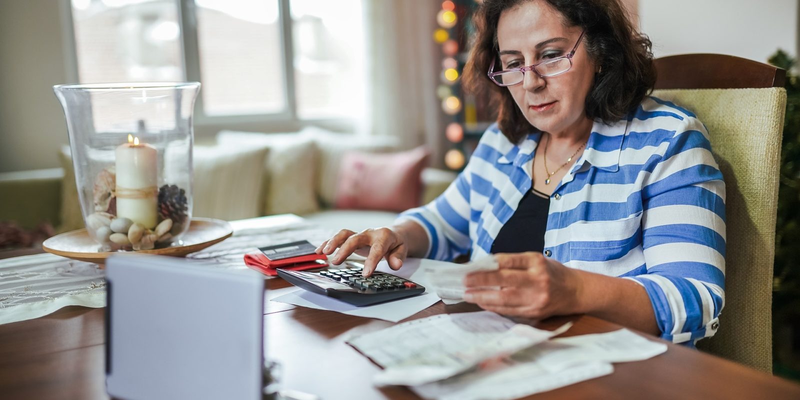 woman in blue striped shirt and glasses sitting at a table covered in receipts and typing on a calculator
