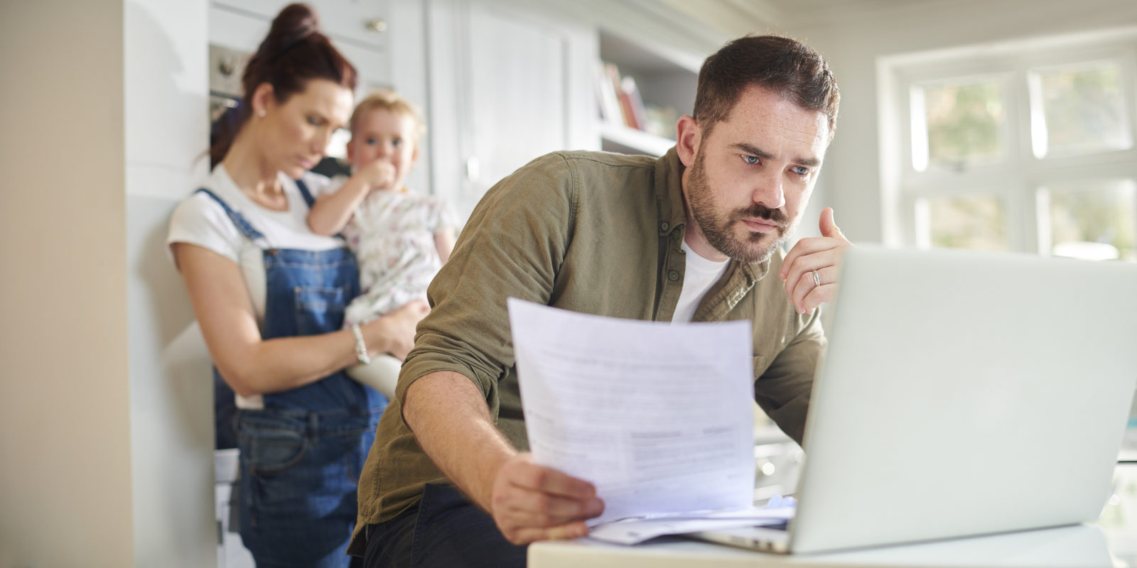 man sits at laptop and holds a piece of paper, looking concerned, while woman in overalls and baby look over his shoulder