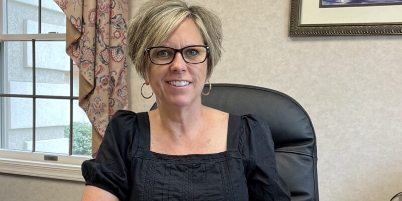 blonde woman in black shirt and glasses sitting in a leather desk chair and smiling