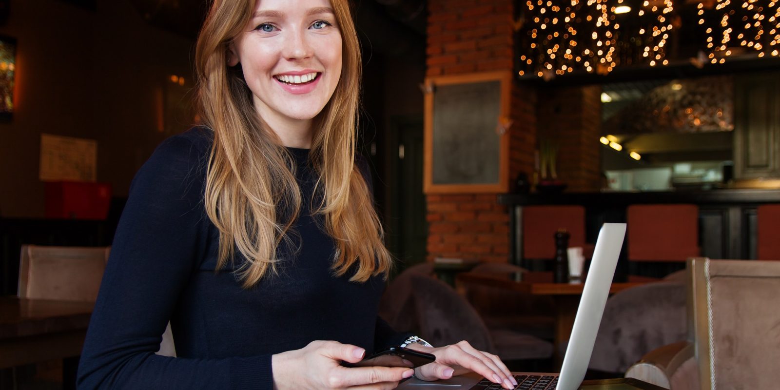 Red haired woman in a black shirt sits at a table in front of a white laptop. Bricks and twinkly lights in the background