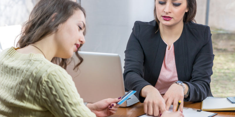 Woman in black blazer explaining something on clipboard to woman in green sweater who is holding a credit card
