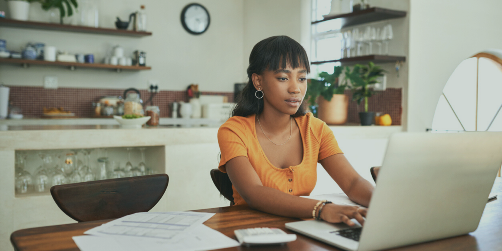 woman in orange shirt sits at a wooden kitchen table and types on a gray laptop
