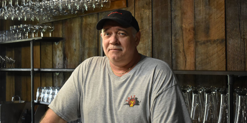 man in a grey t-shirt and a black baseball hat looks at the camera, glassware above him and in the background
