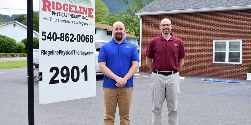 Two men stand in front of a brick building next to a sign that reads 