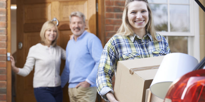 woman in flannel smiling and loading a box into the trunk of a car while parents smile in the background