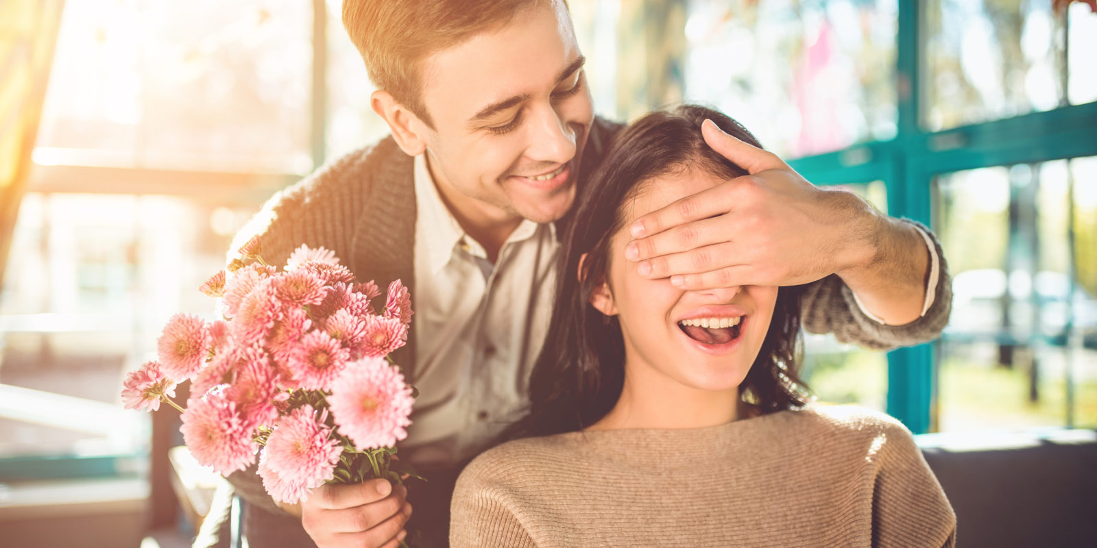 man in a white shirt and gray sweater holds pink flowers and covers the eyes of a woman in a brown sweater